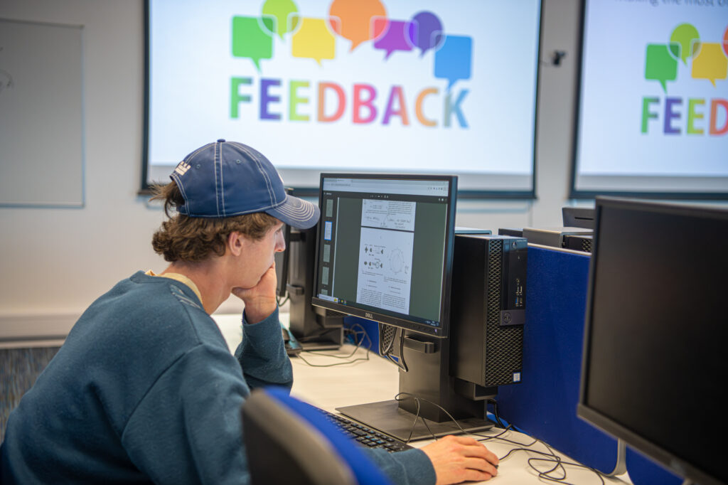 Picture of a student sitting in a computer lab with the word 'feedback' written on the screen behind him.